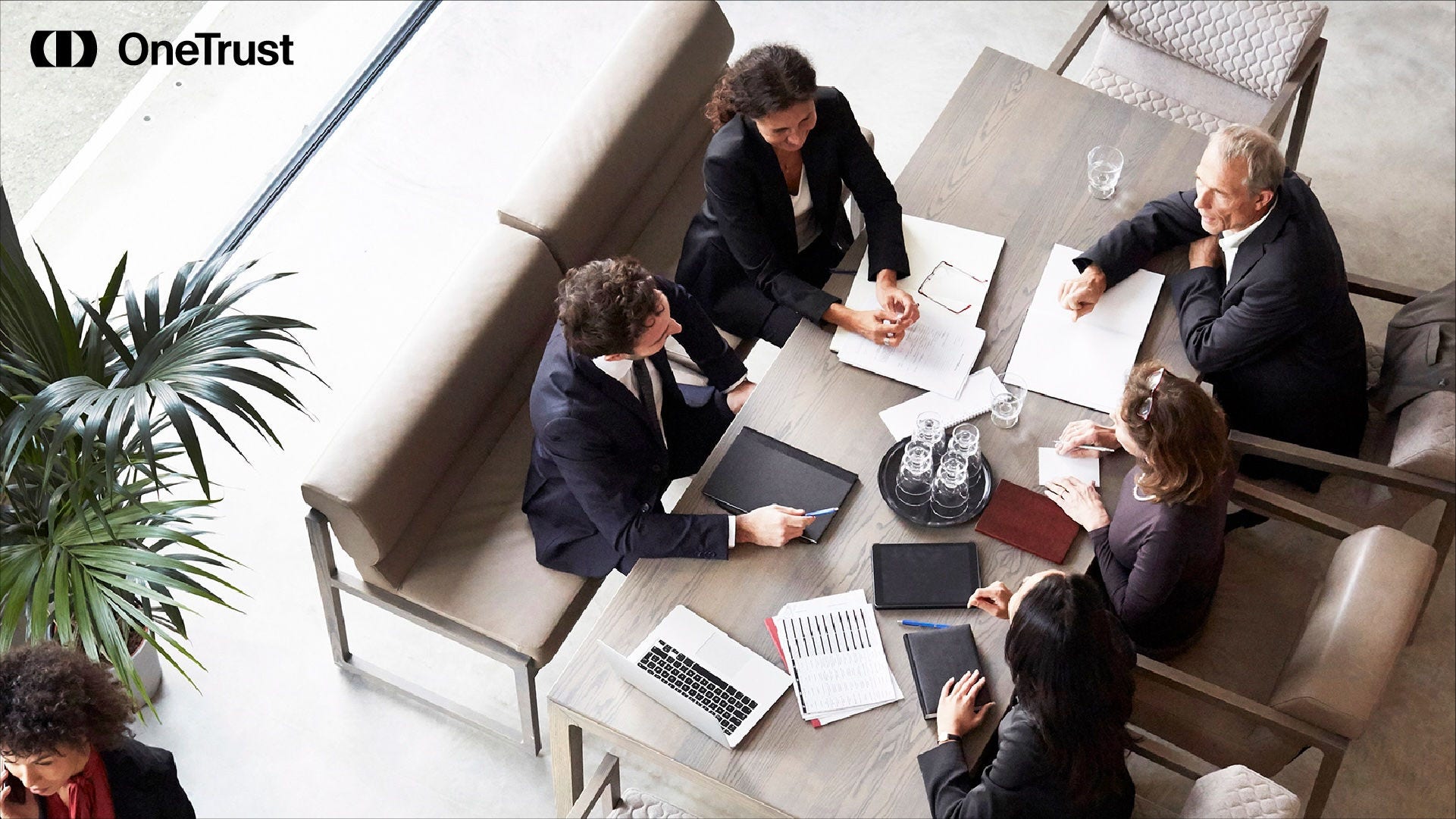 Five professionals meet around a table reviewing documents, laptops, and tablets in a modern office space.