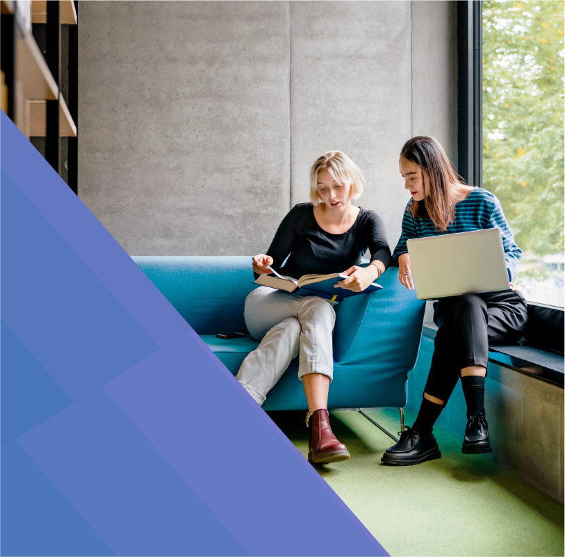 photo of a white woman and an Asian woman in their 20s collaborating on a project. One woman is sitting on a blue couch white the other sits on a low windowsill. 